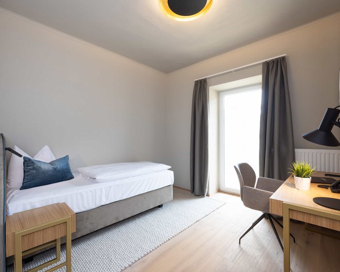 Helles Einzelzimmer mit Schreibtisch, großem Fenster und Bett.