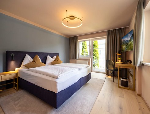 Doppelzimmer mit großem Bett, Schreibtisch und Balkonfenstern.